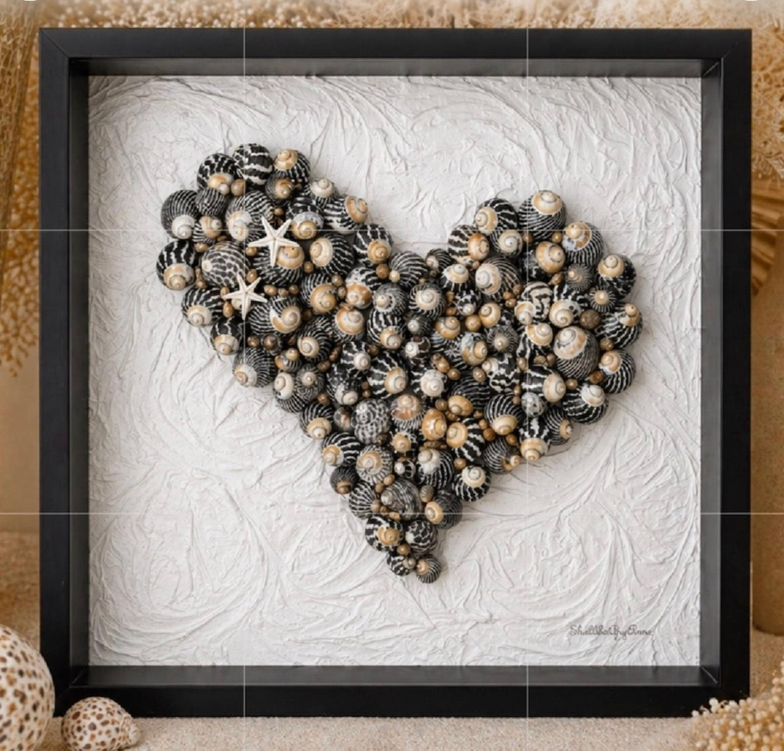 Handmade coastal shell art love heart in black shadow box frame on textured white background, made with zebra sea shells, starfish and gold sea beads