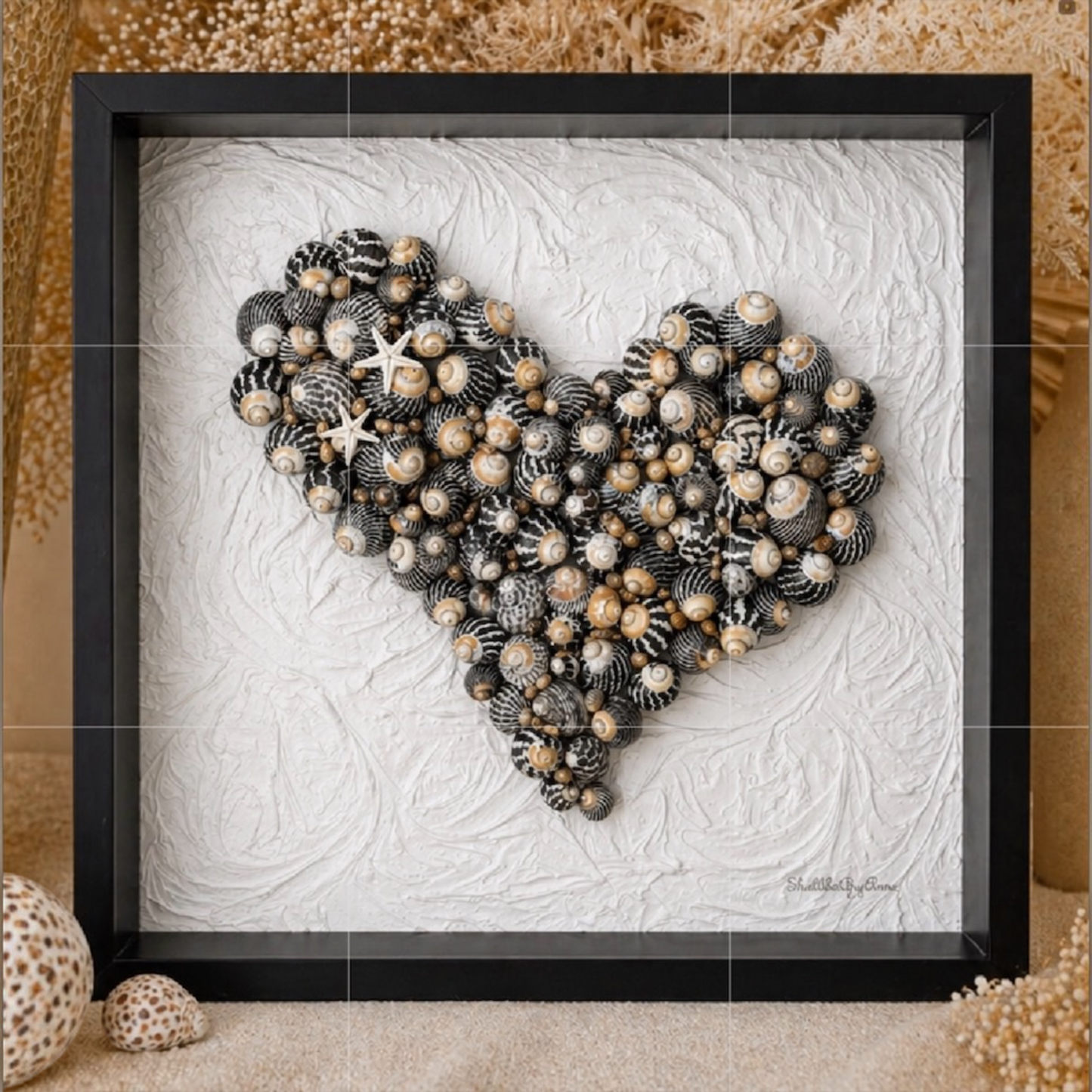 Handmade black and white zebra shell art heart in black shadow box frame styled with coastal decor on sandy background, unique coastal gift